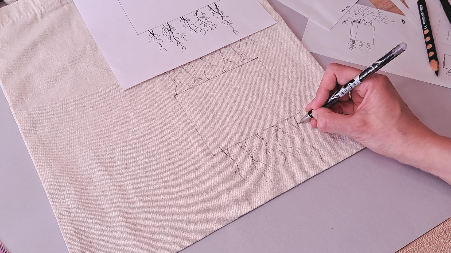 Hand drawing a carrot and root design onto a natural canvas tote bag with an erasable fabric pen, with printed reference sketches visible nearby