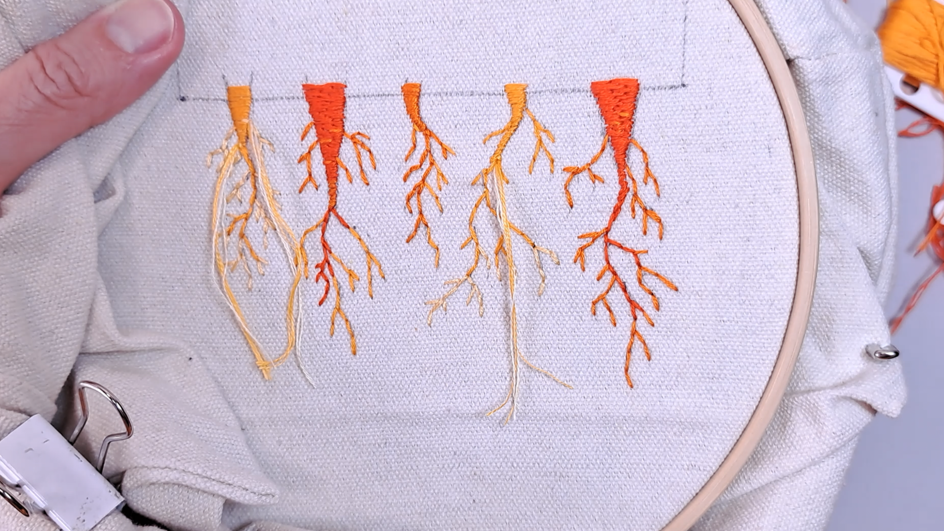 Close-up of hand-embroidered carrot roots in an embroidery hoop, stitched in back stitch using shades of orange, red, and pale yellow, showing varied colour gradients across five carrots